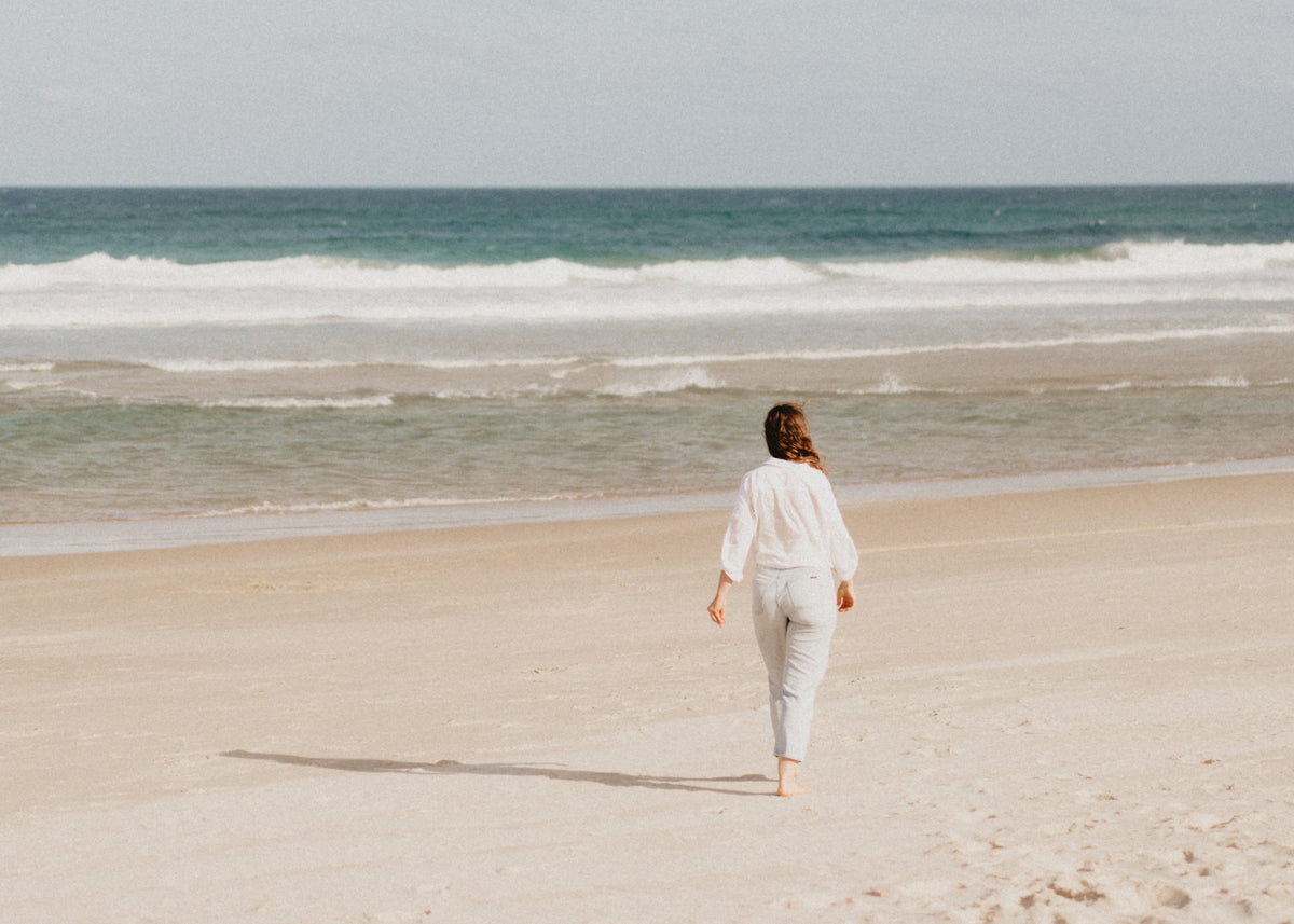 A woman in a white long-sleeve top and light pants walks barefoot on a sandy beach toward the ocean under a pale blue sky.