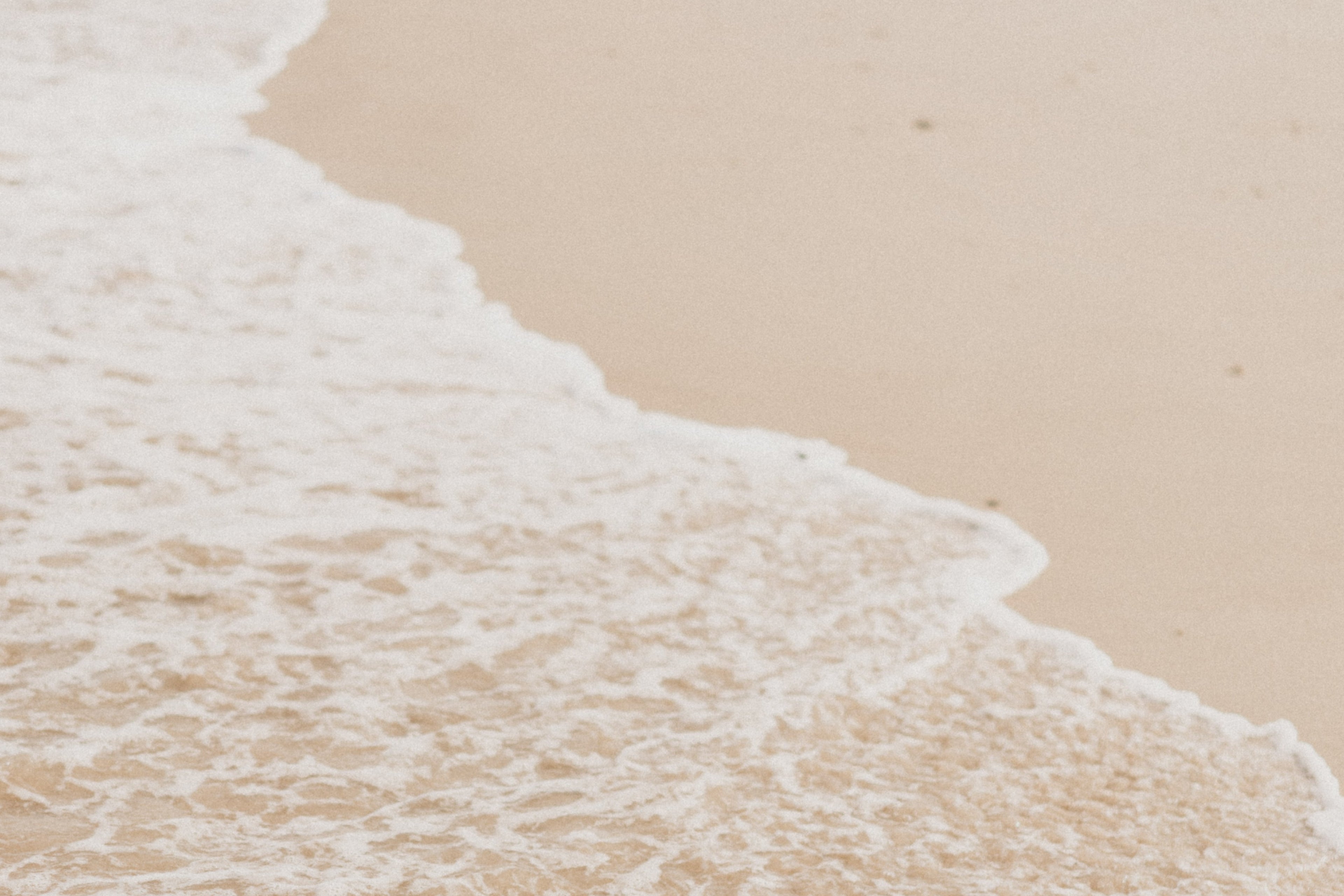 A close-up beach scene showing pale sand with foamy white ocean water washing onto the shore.